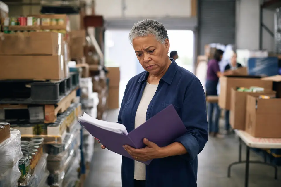 Food bank director reading paperwork in a warehouse aisle between stacked boxes